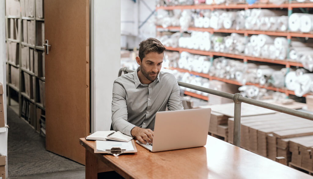 Shipping logistics professional (probably retail) on a laptop with inventory stacked in the background